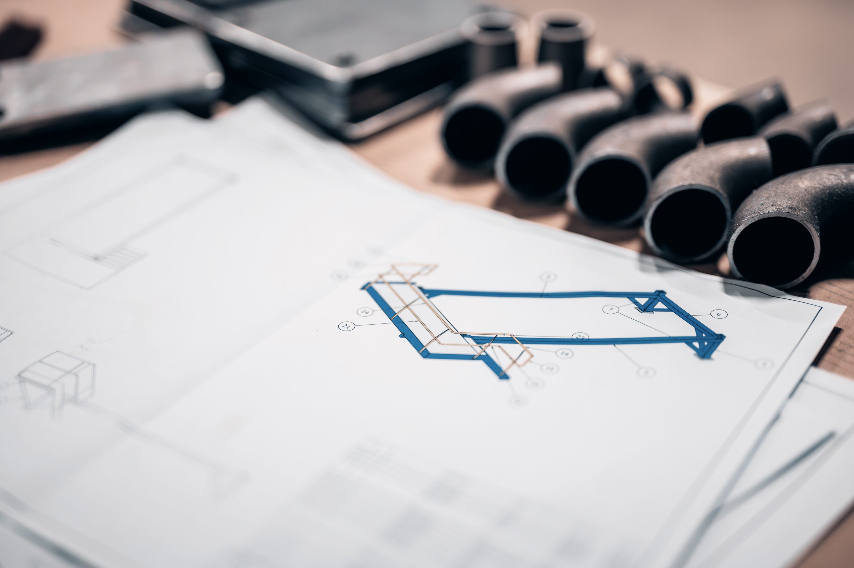 Technische werktekening van een constructie op een werkbank, met metalen bochtstukken en materiaal op de achtergrond. D-Novus
